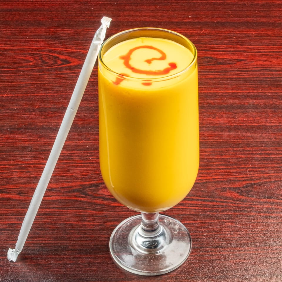 A tall glass of yellow mango lassi with a red swirl on top, next to a white straw.