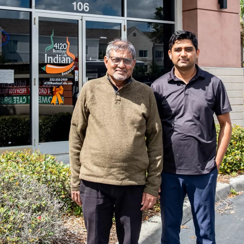 Two men, one older and one younger, stand smiling in front of Bombay Cafe Indian Restaurant.