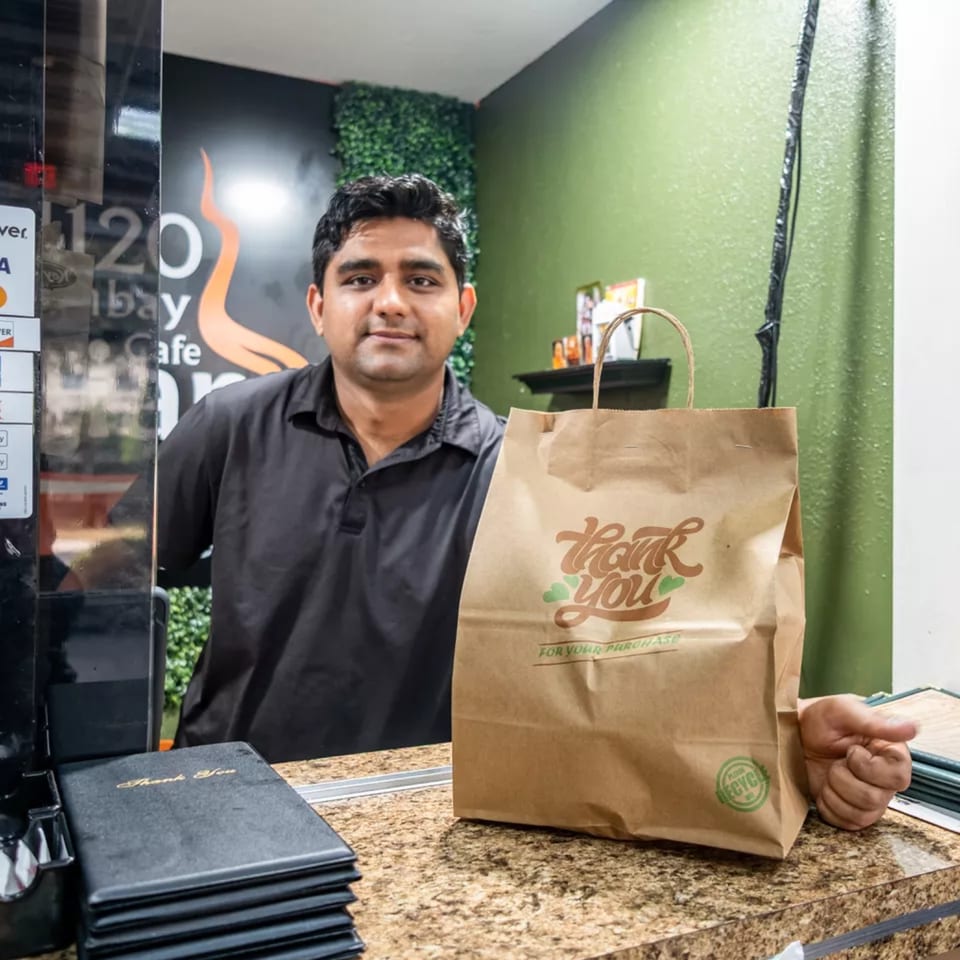 A Bombay Cafe employee stands behind a counter with a "Thank You" takeout bag.