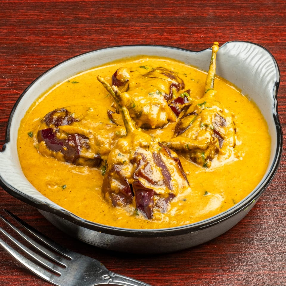 Indian eggplant curry in a white bowl with a fork on a dark red wooden table.
