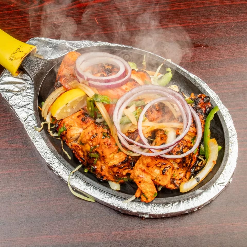Steaming hot chicken tikka sizzler with onions, peppers, and lemon on a dark wooden table.