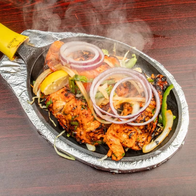 Steaming hot chicken tikka sizzler with onions, peppers, and lemon on a dark wooden table.