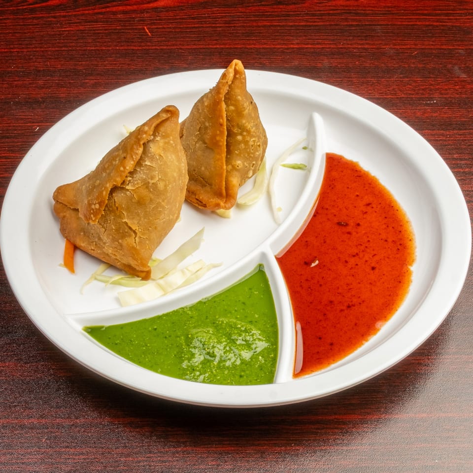 Two golden-brown samosas with shredded cabbage, served with green and red chutneys on a white plate.