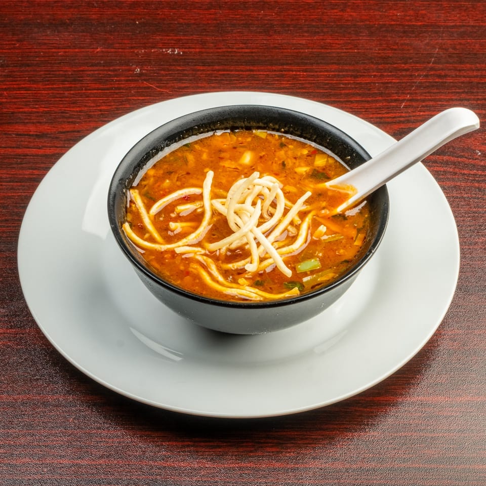 A black bowl of hot and sour soup with noodles and a white spoon on a white plate.