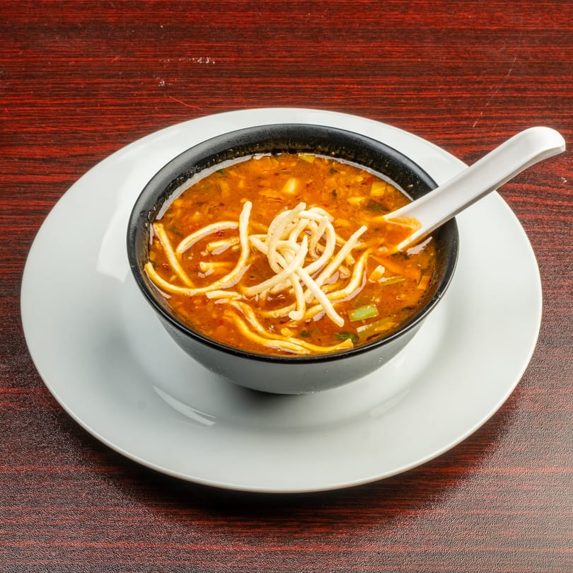 A black bowl of hot and sour soup with noodles and a white spoon on a white plate.