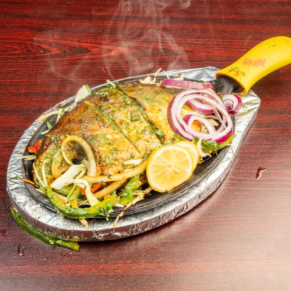 Steaming whole fish, garnished with onions, peppers, and lemon slices, served on a hot platter.
