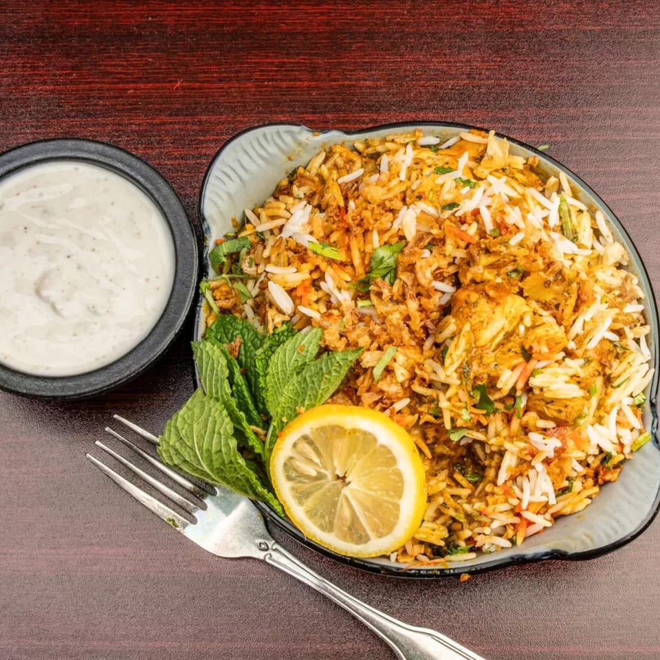 Chicken biryani with mint, lemon, and a side of raita on a dark wooden table.