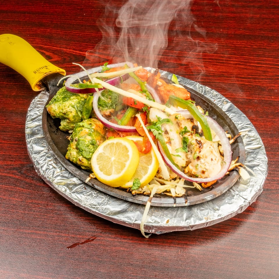 Steaming Indian sizzler platter with chicken, vegetables, and lemon slices on a dark wooden table.
