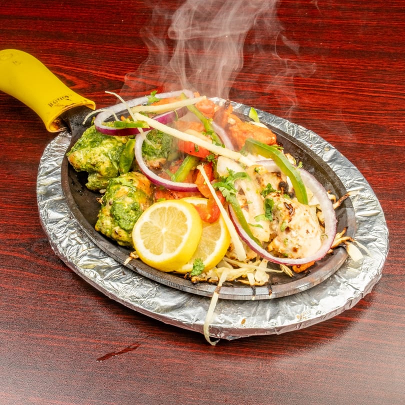 Steaming Indian sizzler platter with chicken, vegetables, and lemon slices on a dark wooden table.